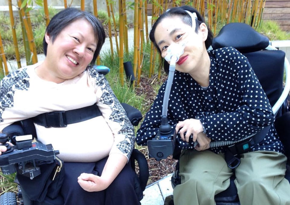 Two smiling Asian women in electric wheelchairs, with the characteristics of spinal atrophy disorder.