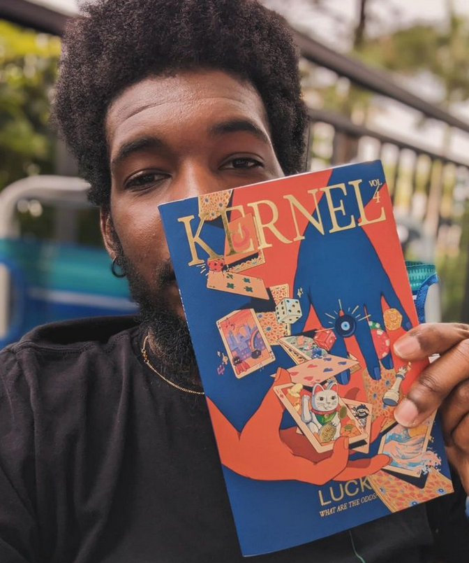 Myself sitting outside holding a copy of Reboot’s fourth volume of Kernel, titled “Luck”.