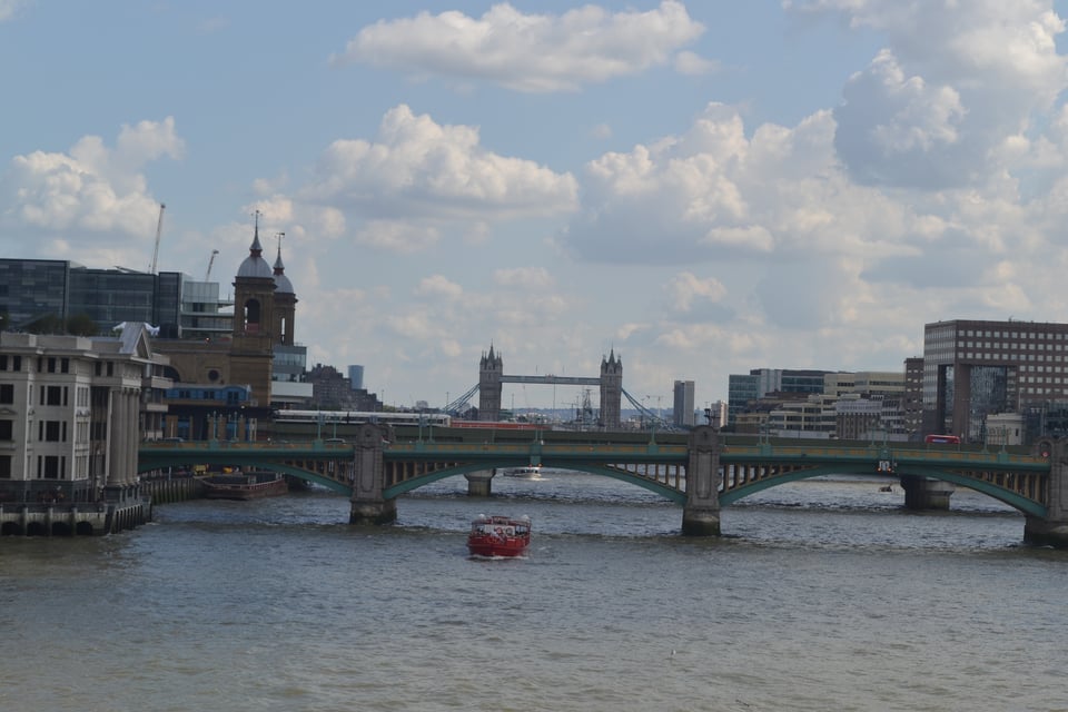A view down a river from a bridge. Two low bridges in the foreground, and London's tower bridge in the background. Various buildings on the banks and a red boat under the nearest bridge.