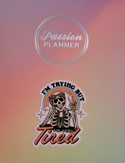 cover of a rainbow passion planner with a sticker. The sticker is a skeleton with sunglasses under a sparkling rainbow. The sticker says,