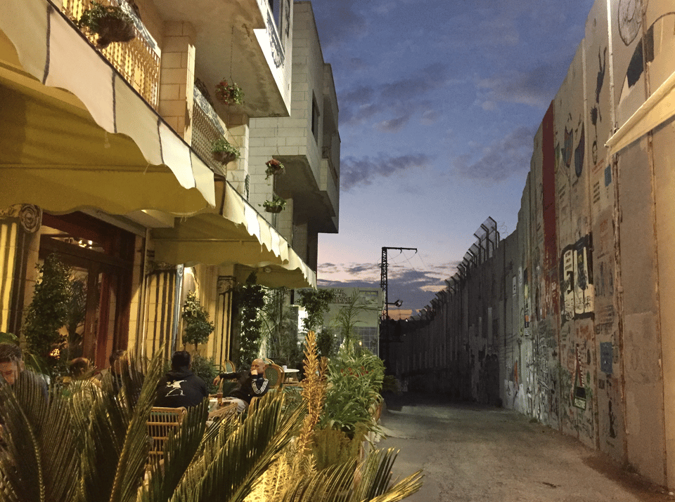 Photograph depicting a narrow alley beside a warmly lit building with balconies, along with outdoor tables and chairs, and many potted plants. On the other side of the alley is a tall concrete wall that appears to be covered with graffiti. The wall is close to the building, and is partly topped with wire fencing. The sky is at dusk.