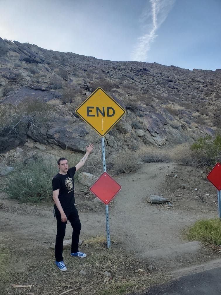 A man in black points at a road sign that says END. Immediately behind the sign is a large mountain