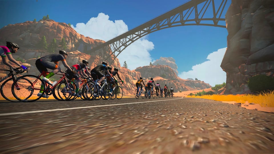 Screenshot of virtual cyclists onscreen racing in the Zwift online cyling-gaming platform. They are riding on a virtual desert road, heading underneath an arched metal bridge that spans a canyon