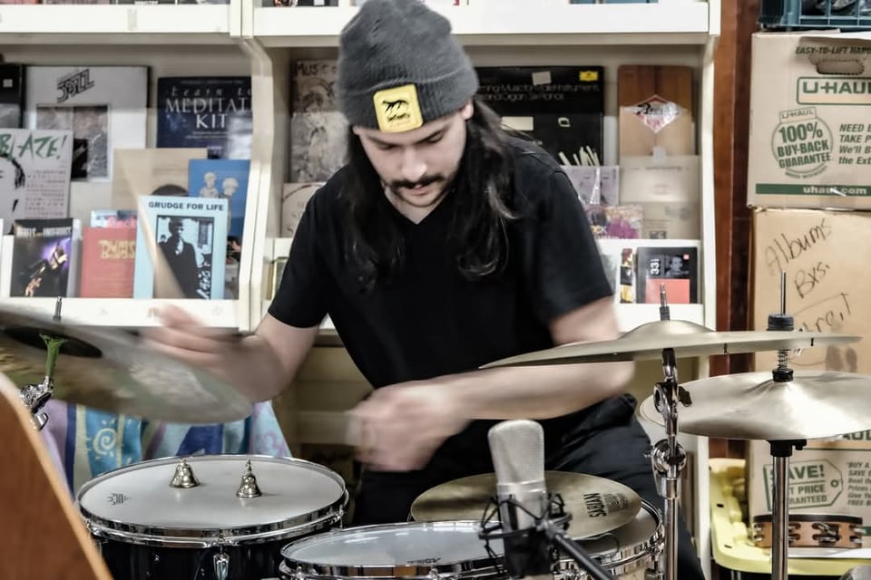 A person with a mustache and shoulder-length hair, wearing a beanie and a black t-shirt, plays a drum kit.