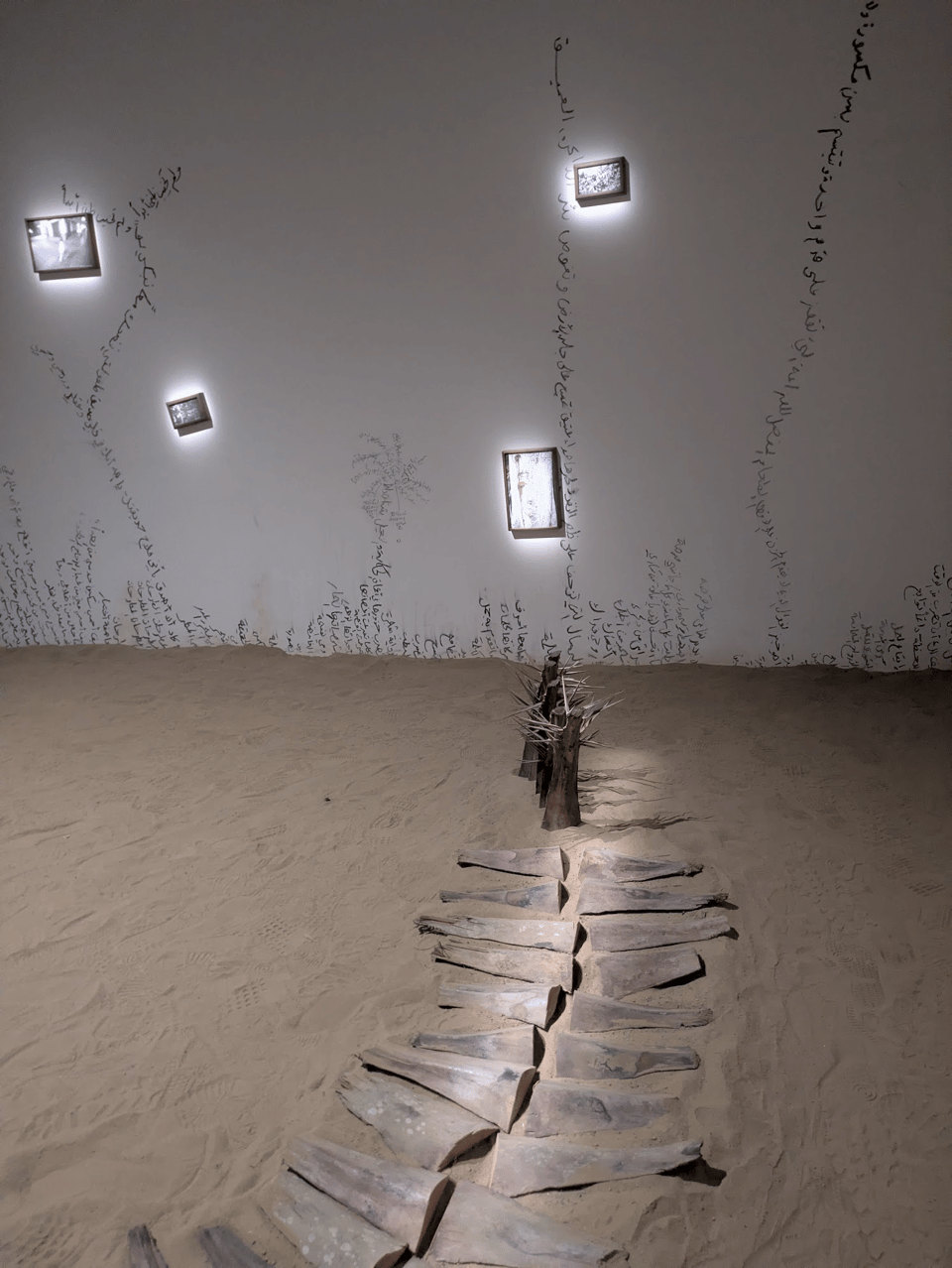 A photo of the art exhibit "Like Tea Leaves in Water," which is described in the below article. This particular photo is focused on wedges of palm wood that sit on sand in a room - the wedges are arranged in a way that suggest a path or a spine. They lead to a series of upright pieces of wood with spikes sticking out of them. The wall in the background has Arabic charcoal writing that evoke plants growing up the wall. Some of the writing "blooms" into photographs that are not clearly visible.