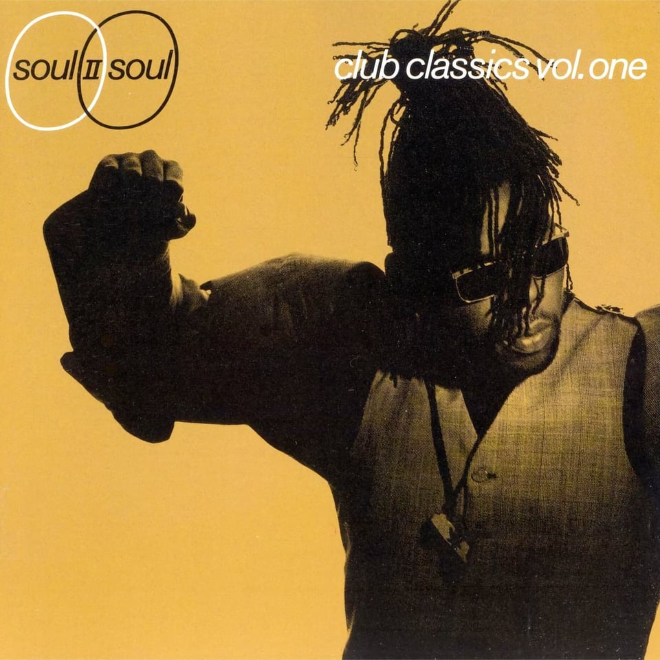 A yellow-tinted black and white photo of Soul II Soul bandleader Jazzie B, from the waist up. He's a Black man wearing a vest and a necklace with Africa on it. He has dreadlocks, and they're mostly tied up out of is face save for a few strands falling across his face. He is the anti-Don Henley. He appears to be pumping his fist. He looks to the right with a blank expression.