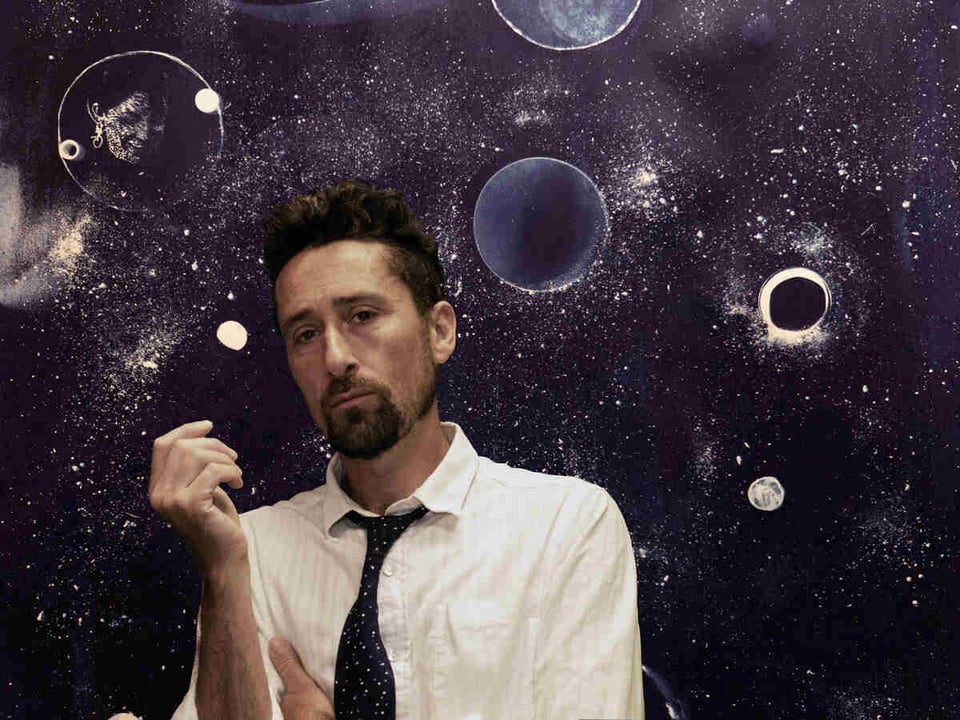 A man with a beard and upswept hair wears a white shirt and dark necktie as he stands in front a backgrop that seems to show planets or celestial objects. In one object above him to his right there's a woman's face.