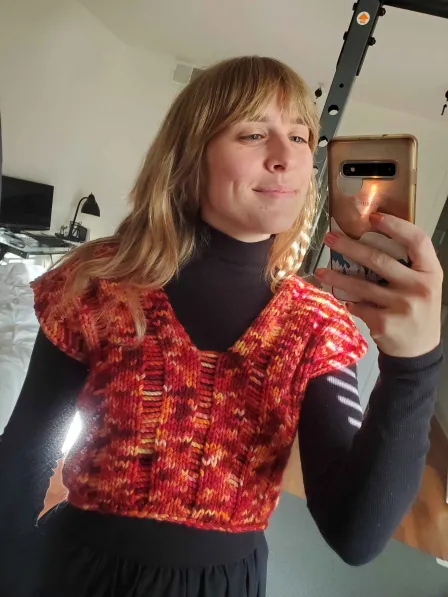 A person with long blonde hair wears a red, yellow, pink and orange top that's been knit with a v neckline and short sleeves. There's a pleating pattern, I think? I don't know a lot about knitting, I just think it's cool.