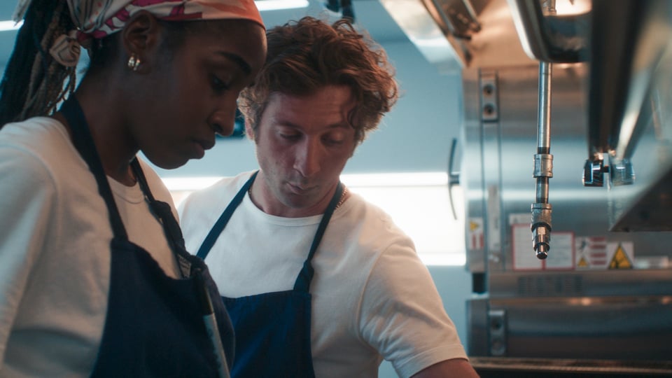 Sidney and Carmen are in the restaurant's kitchen, surrounded by stainless steel appliances, wearing aprons, gazing down at something Sydney has cooked up.