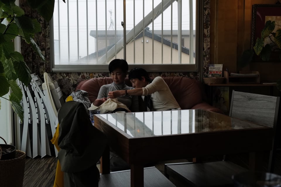 Two women seated together on a worn pink sofa inside a homely Japanese cafe, one tending to a small infant, tiled rooftops visible through louvred windows behind them
