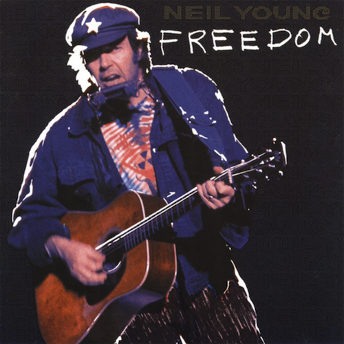 A color photgraph of Neil Young, a white man in a blue jacket, blue cap with a white star on it, and tie-dyed shirt with a radiation symbol on it. He is singing. He exhibits the most "cranky uncle" energy in the universe. He's playing acoustic guitar and has one of those harmonica holders on his neck. It looks like it was shot in concert.