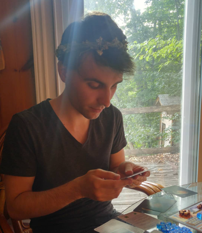 Kurt (the author) looks at a card held in hand. Behind him is a forest through a glass door. On his head, he wears a crown made of broadleaf plantain and clover.