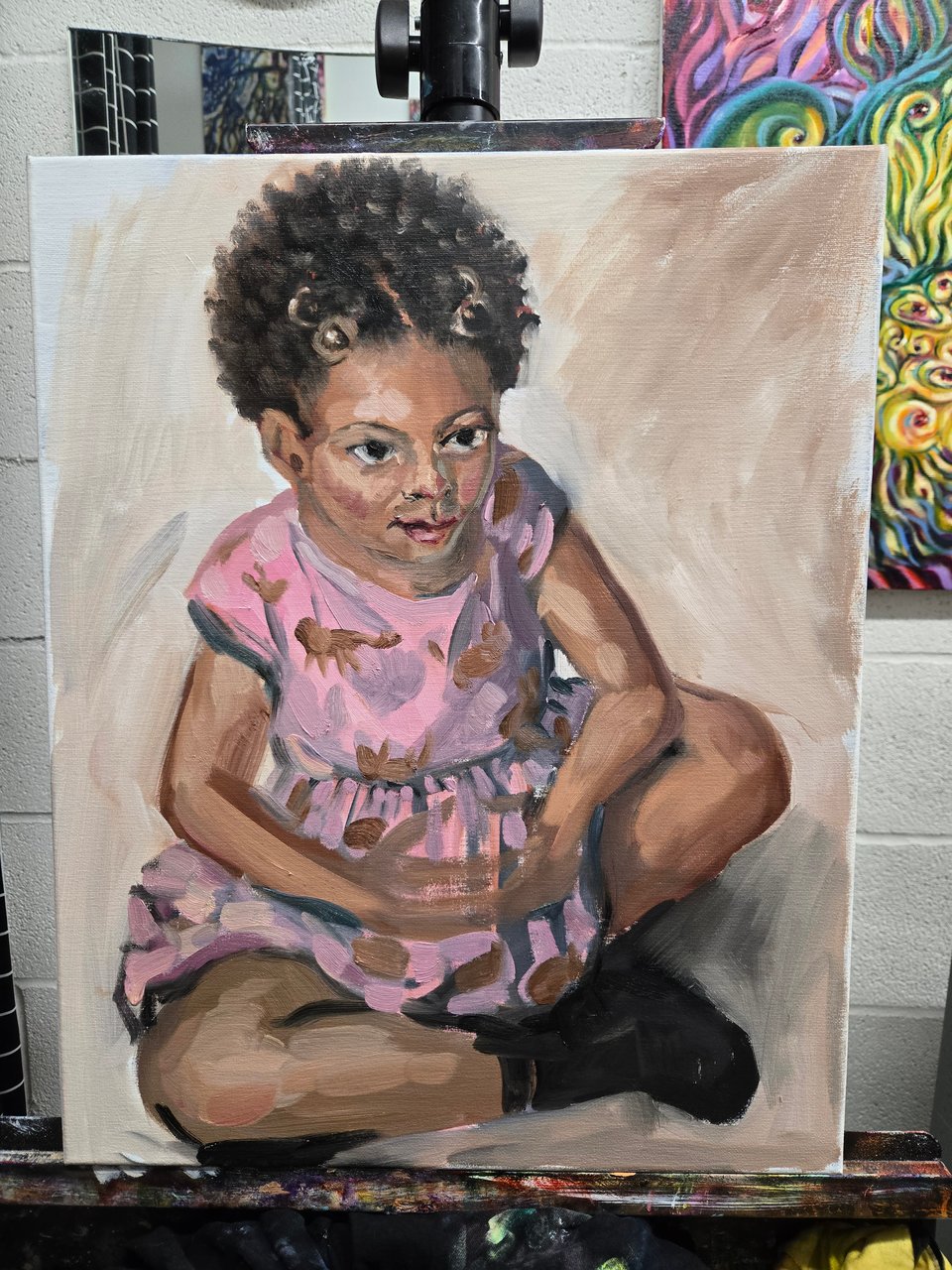 photo of an unfinished painting on the easel, of a bronze skinned preschooler seated on the ground in a pink dress and black shoes, hair in two bubble-tie puffs, hands steepled