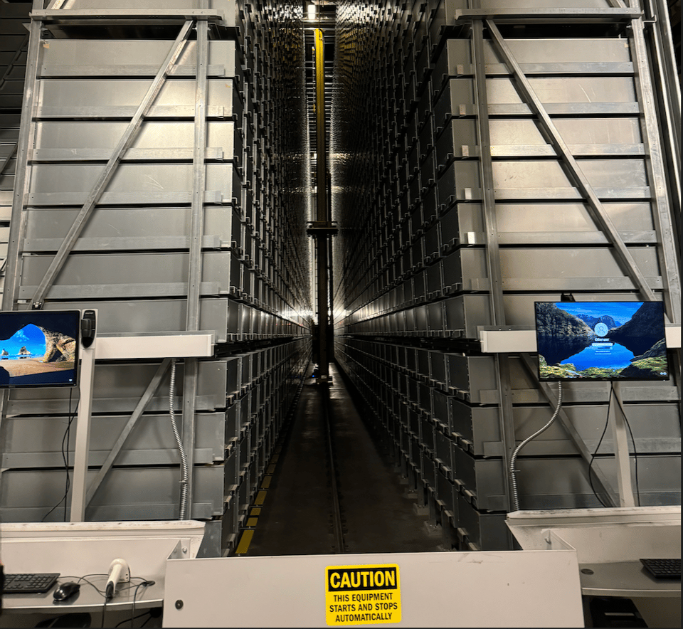 The yellow vertical arm of the book selection robot recedes between two giant metal walls, with a computer screen on each side.