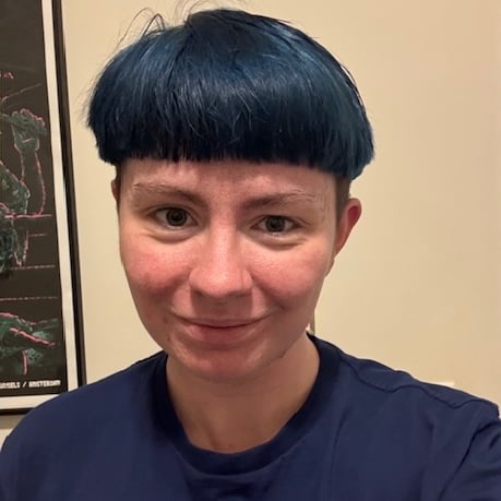 A white person with blue hair in a blue shirt smiles at the camera against an off-white background.