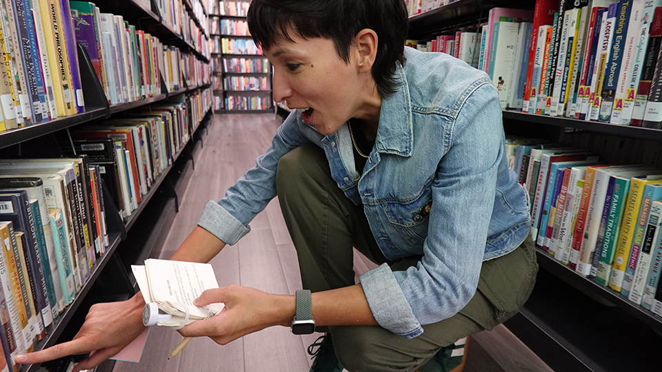 A woman in a denim jacket is delighted to find something unique in the library shelves.