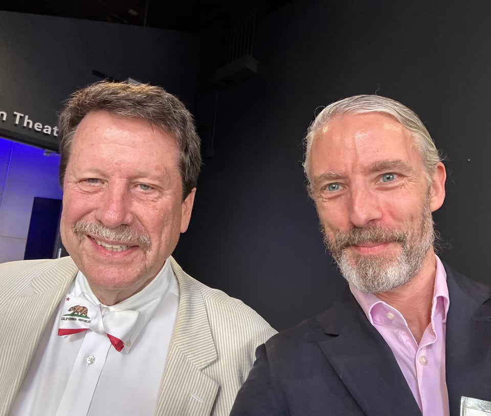Photo of two white man in suits; the man on the left is wearing a beige & white seersucker, with a white shirt and a bow-tie of the flag of California; the guy on the right is me, and I should've trimmed my beard more before this trip