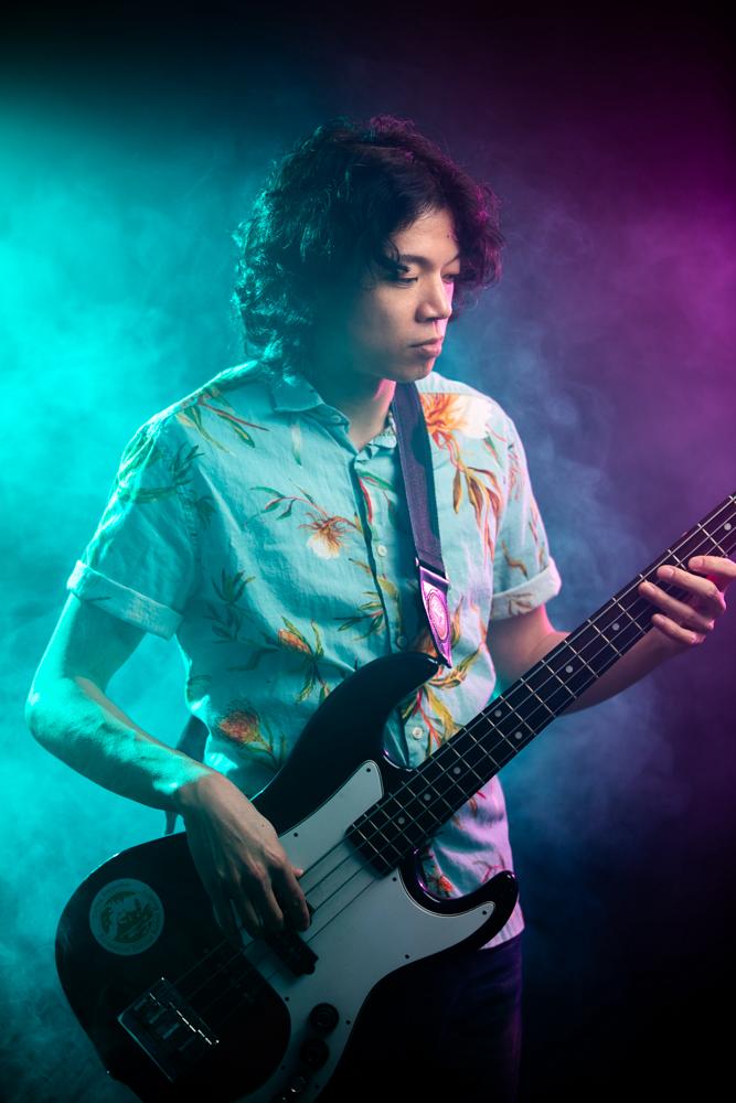 A man with shaggy hair wearing a floral-print short-sleeve shirt plays a bass guitar.