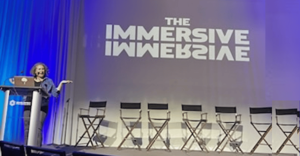 A woman with curly hair presenting on a stage with a screen that says the Immersive Immersive