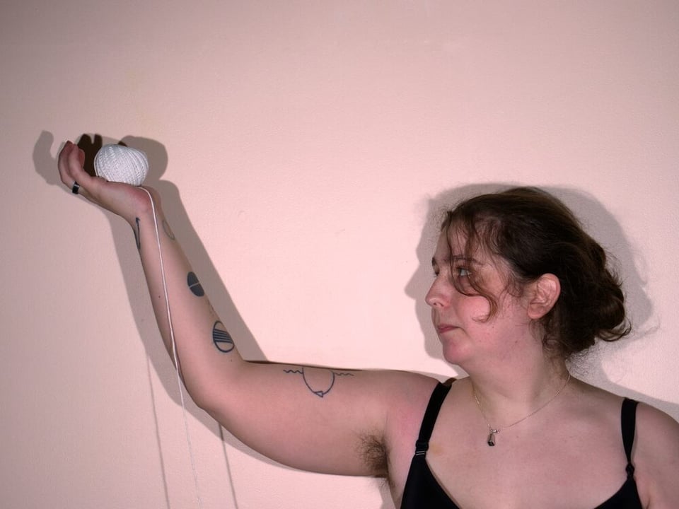 A woman shown from the upper chest up stands on the right side of the frame, looking to her right with her arm upraised and a ball of twin in her open palm.