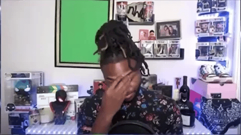 young man with dreadlocks is sitting at a desk in front of a green screen. He has his hand on his face, looking disappointed or upset.