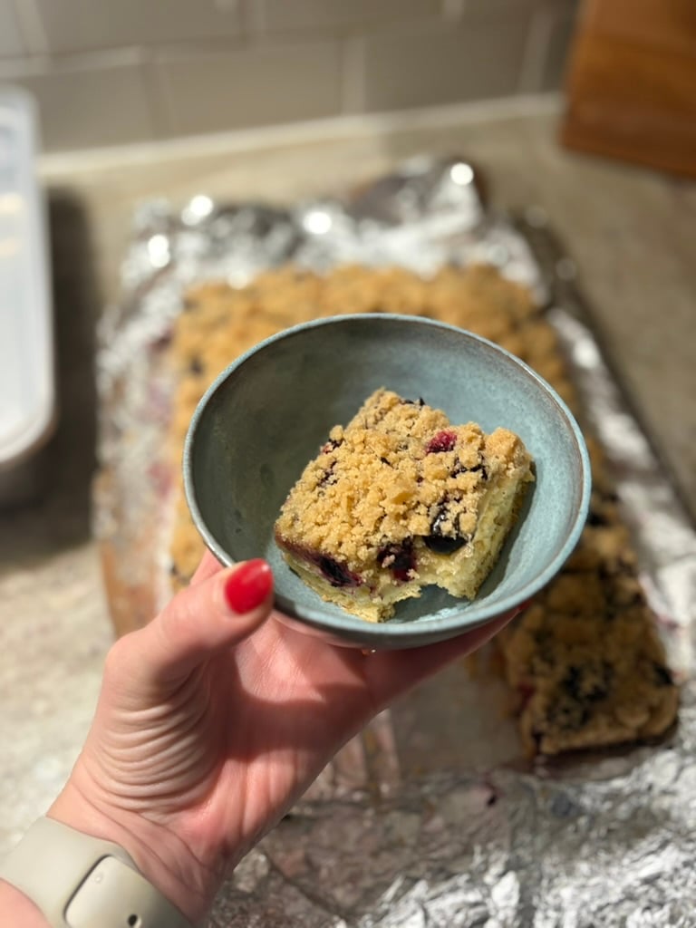 A slab of blueberry streusel in the background with a small turquoise bowl containing a square piece of streusel in the foreground.