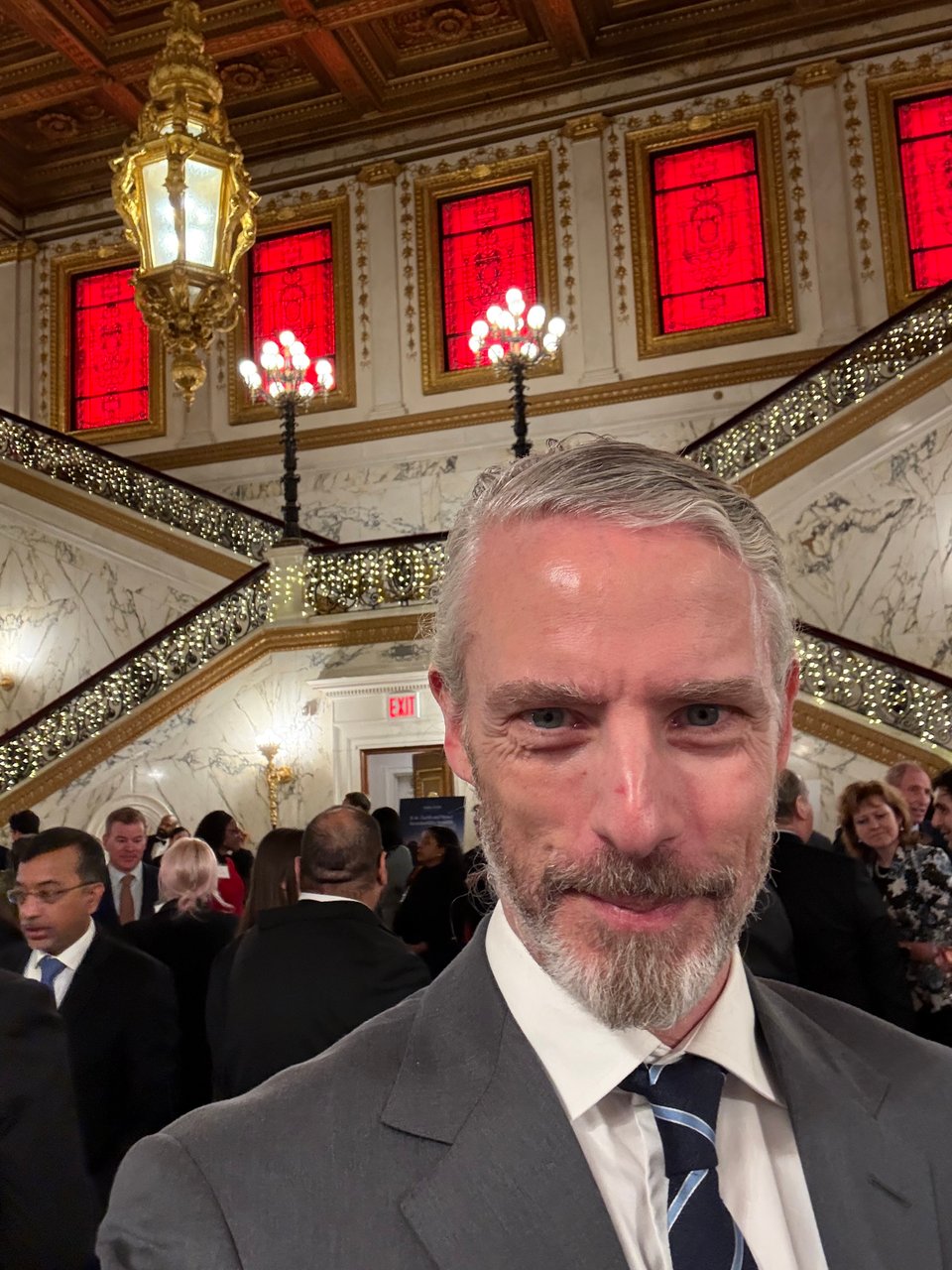 photo of white man in gray suit, white dress shirt and navy tie with cornsilk stripes, standing in the reception area of The Metropolitan Club in NYC, with many people in dark suits behind him. The walls are marble, and the railings of the stairs in the background have white holiday lights. Red stained glass windows are a floor above