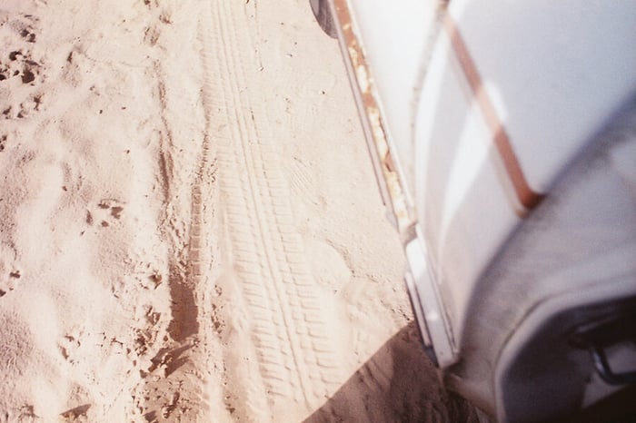A over-exposed, summer sun-bright photo of a tire track in the sand. On the right of the image is the out-of-focus body of a rusting old white pickup, as though the photographer were leaning against it.