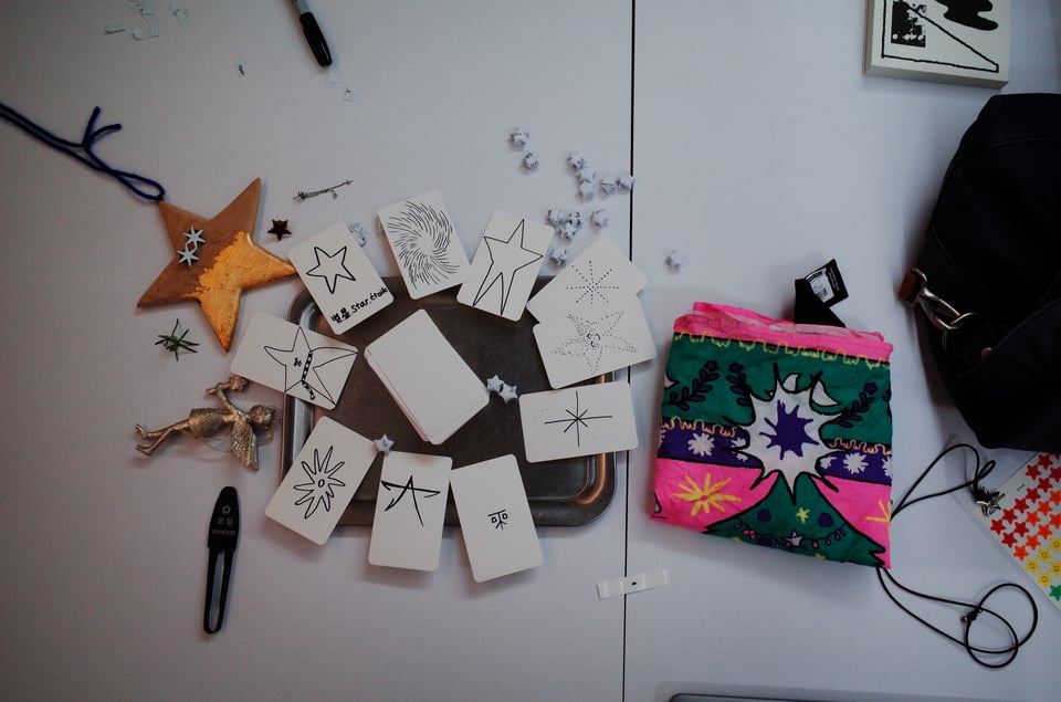 An aerial view of a white table shows several star shaped objects — pins, ceramic stars, stickers, a patterned handkerchief – alongside drawings of these star-shaped objects on index cards.