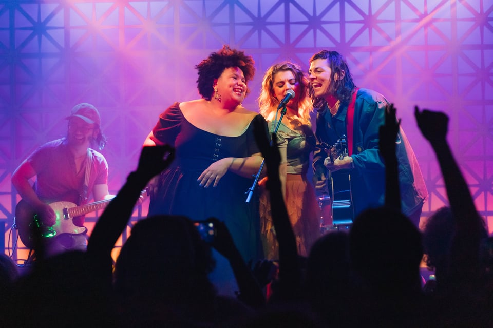 Regina, Sydney, and Adrian sign into a mic. The crowd raises their arms, and a guitarist behind them smiles while playing.