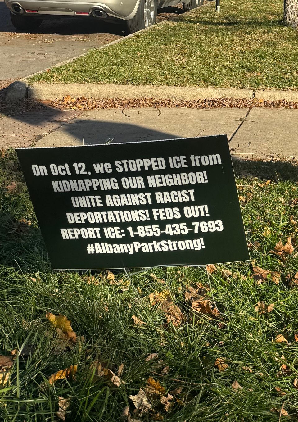 A black yard sign with white text: On Oct 12, we stopped ICE from kidnapping our neighbor! Unite against racist deportations! Feds out! Report ICE: 1-855-435-7693 #AlbanyParkStrong
