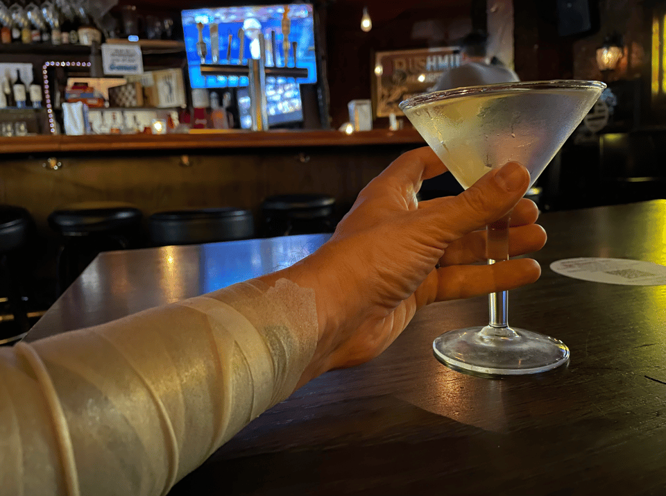 A person with a bandaged arm holds a chilled martini in a wood-filled bar