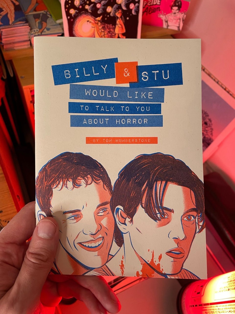 A hand holding a zine, "Billy & Stu would like to talk to you about horror by Tom Humberstone", featuring art of Billy and Stu from Scream