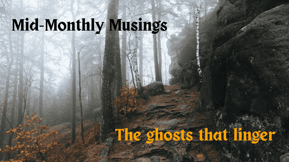 A photo of a foggy, misty woods in late autumn. Some low shrubs retain orange leaves. There is a path at right, moss-covered, made mostly of barely-visible roots and packed dirt, winding up next to a wall of stone. The words "Mid-Monthly Musings" is at top left in black, while "The ghosts that linger" in ochre-orange are at bottom right.