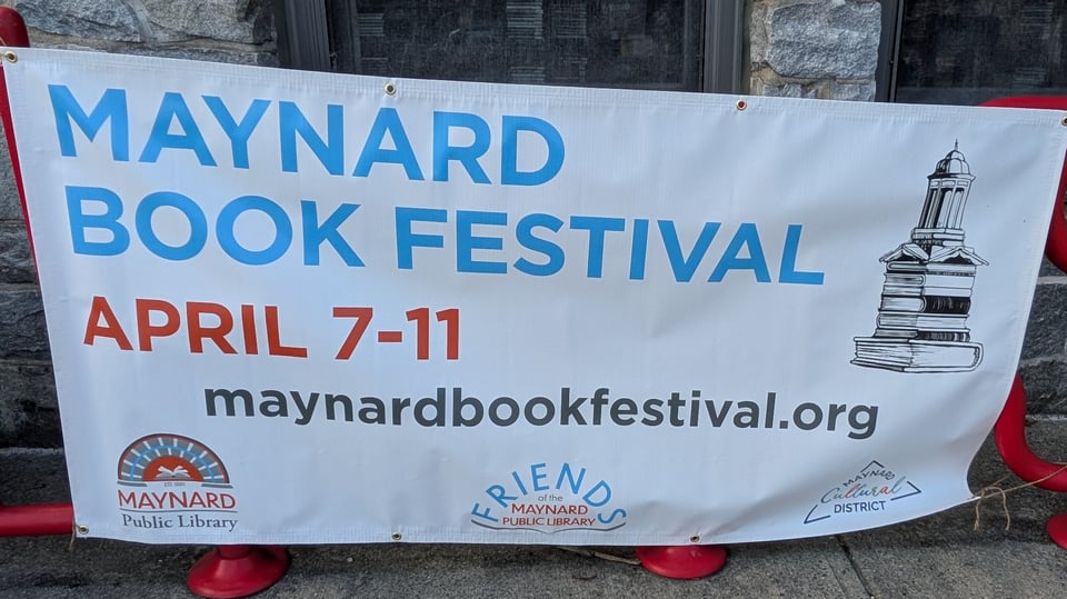 A banner hangs outside the Maynard Public Library. It reads Maynard Book Festival, April 7-11, maynardbookfestival.org. It has the logos of the Library, the Friends of the Library, and Maynard Cultural District on it.