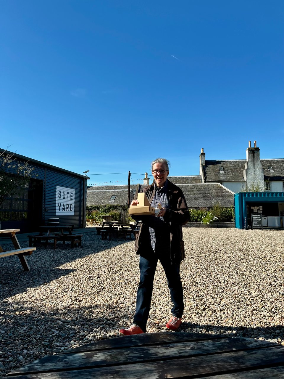 Rowan Ambrose is walking across a pebbly courtyard carrying small cardboard boxes containing food. There's a mixture of very old stone buildings and brand new warehouse style buildings around the edges of the courtyard. The sky is a vivid and cloudless blue.