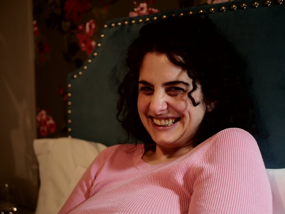 A portrait of a smiling Kat, wearing a pink sweater against a green headboard