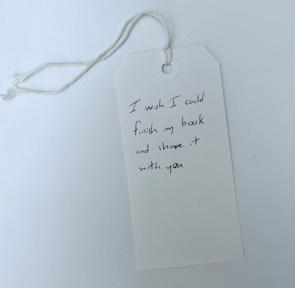 photo of a paper card with the handwritten words, "I wish I could finish my book and share it with you" in ballpoint pen; the card has a reinforced hole-punch and a string running through that