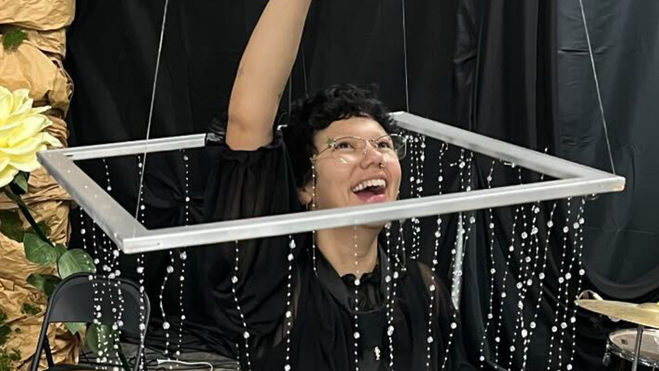 A young person smiling and raising their arm happily whil encircled by a beaded curtain.