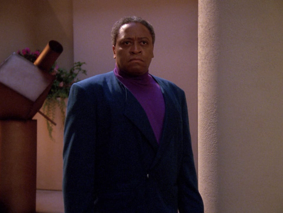 Ron Canada as Martin Benbeck. He is wearing a purple shirt with a blue blazer and appears upset or angry.