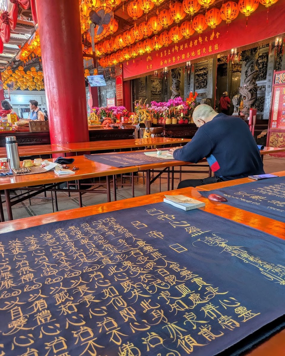 A table inside a brightly lit temple that has blue parchment on it. The blue parchment is filled with beautiful calligraphy in Mandarin, which is a heart sutra (呂祖心經). In the background is a second table with more parchment and a man bent over it currently writing. Above are many lit red paper lanterns, and in the background are colorful flowers - the whole scene is vibrant with color..