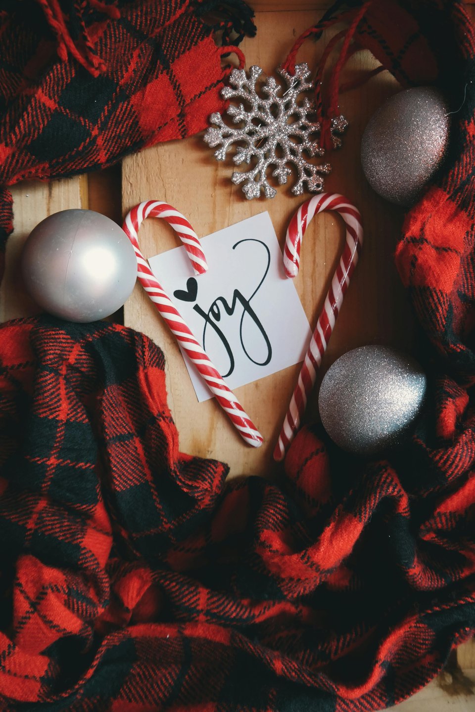 Stock image is of red plaid flanne surrrounding some silver roundornaments and candycanes. In the center of hte image is a piece of paper with the word "joy" written in calligraphy.