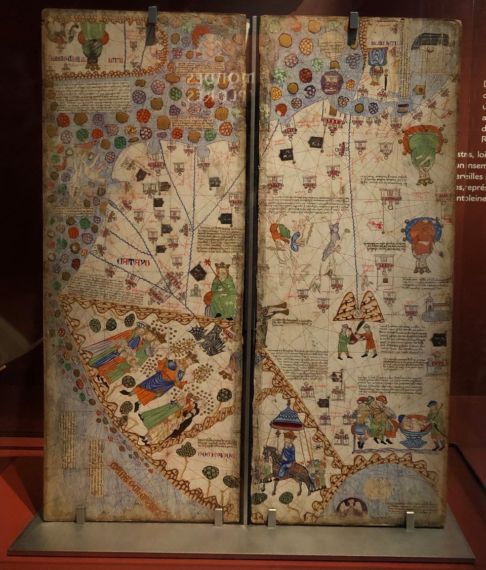 A brightly-coloured, richly-illustrated medieval world map on a vertical stand. The illustrations include kings in varied costumes, people fighting giant cranes, and multi-coloured islands.