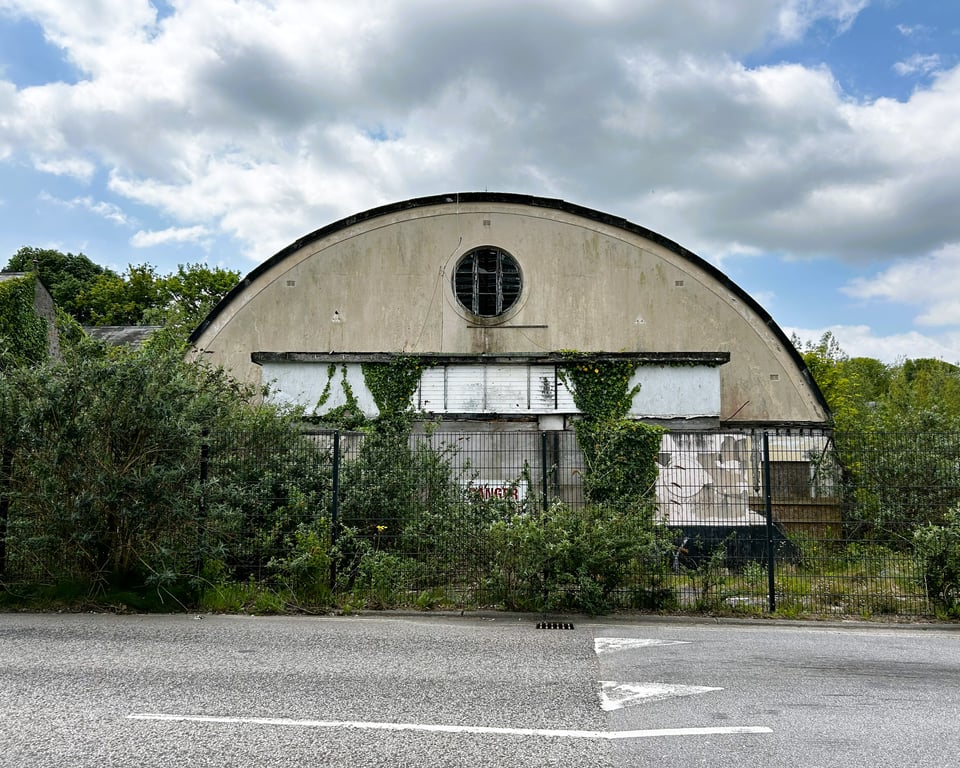 A building with a curved roof that goes all the way to the ground forming a semi-circle. The flat end has been filled with blockwork apart from a circular window that is broken. the building is half obscured by vegetation, and is behind a fence.