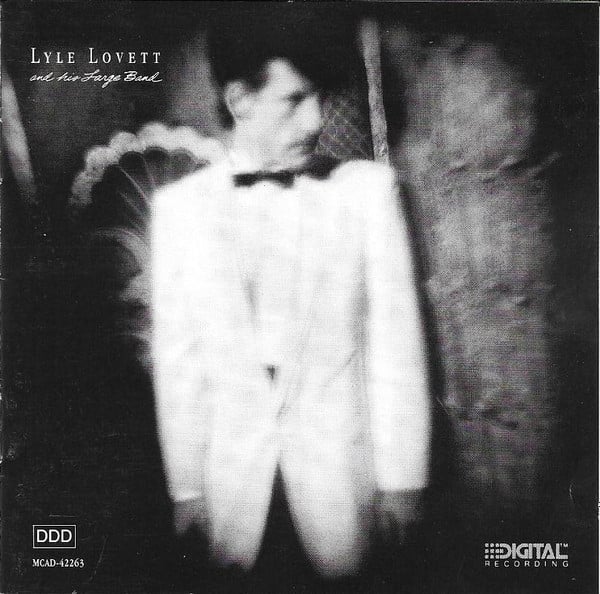 A blurry black and white photo of Lyle Lovett, a white man with a long face and big floofy dark hair that dips over towards the front of his face. He's wearing a big white suit and dark tie that makes him look like a bandleader from the 20s or something. He looks to the right at something out of frame.
