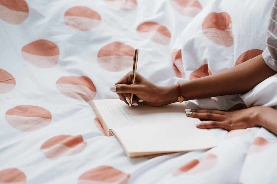 Image shows a pair of hands writing in a notebook while the writer is presumably laying in bed (face not shown).