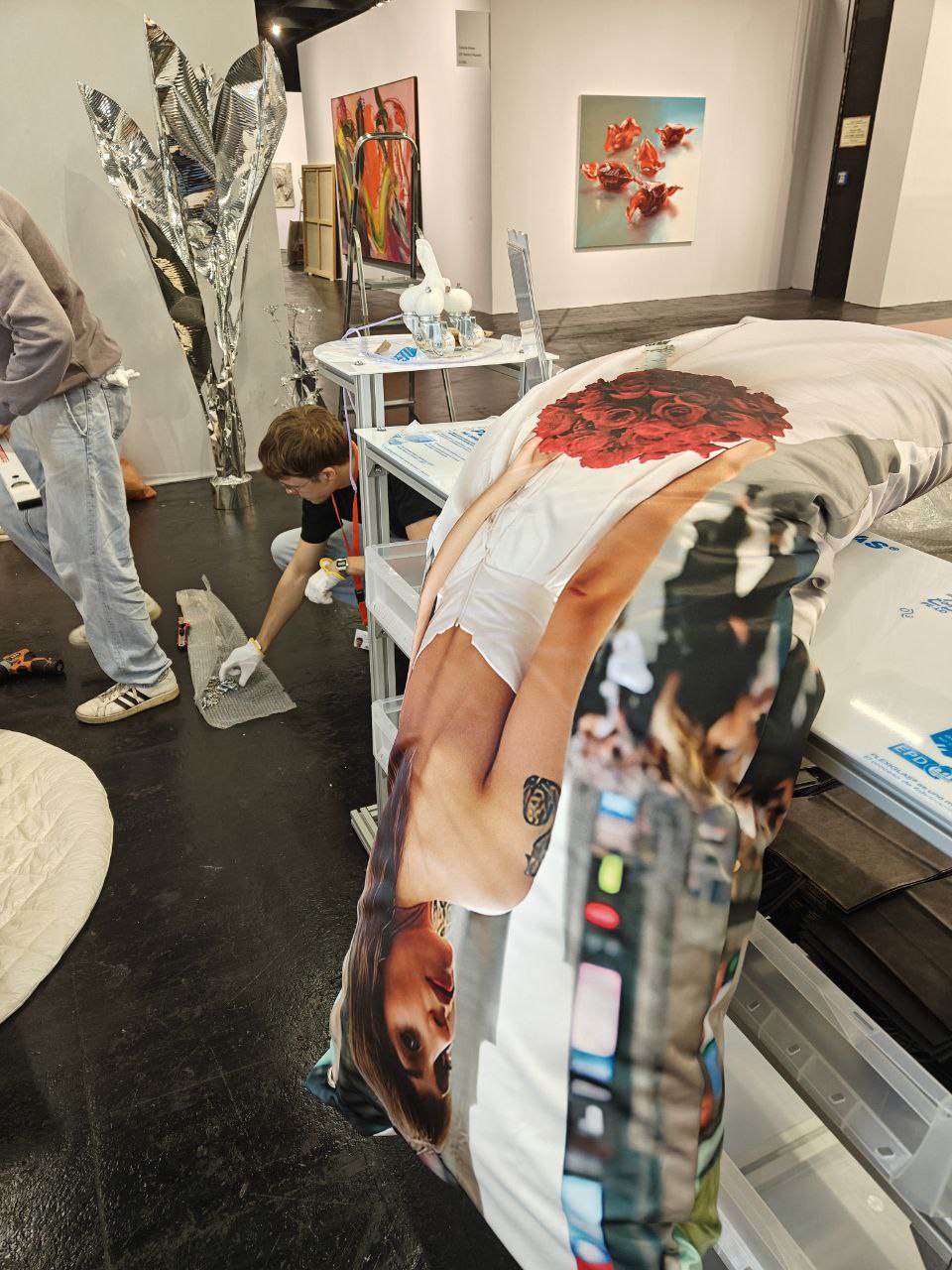The art fair booth midway through installation. A dakimakura of Sarah in a wedding dress is flopped over a white plastic and aluminum display counter. There is still protective wrapping on the table. Towards the back you can see two men, both art handlers wearing white gloves, unpacking things from bubblewrap.