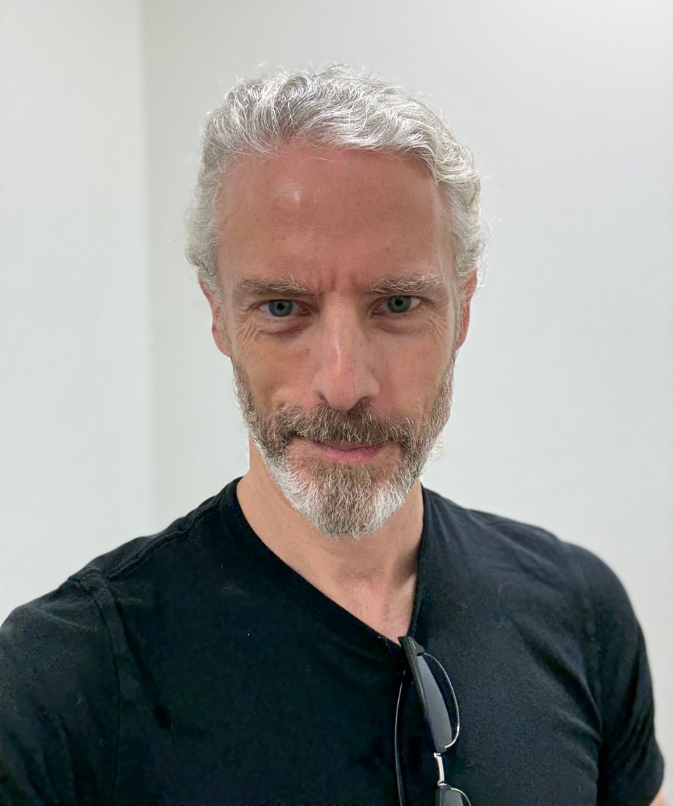 photo of white man with trim beard in a black T-shirt with sunglasses hanging from the neck. The walls behind him are white, as is his hair, which is tied back. He has a slight smirk, and is about to get the results of a blood test