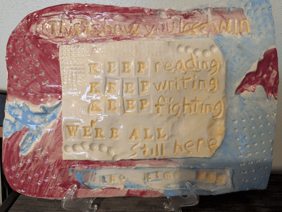 A clay plaque with the following quote on the top and bottom: This is how you lose win the time war. Lose is crossed out. In the middle, it reads: Keep reading, keep writing, keep fighting, we're all still here.