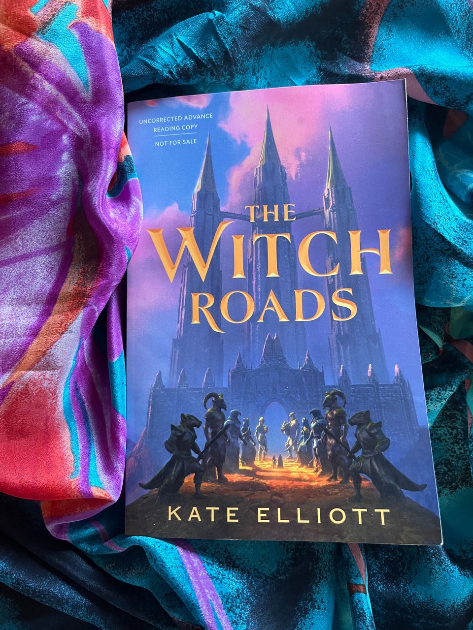 Nestled on a green, orange, and purple scarf lies a copy of THE WITCH ROADS, Kate Elliott. The cover depicts three tall towers in the background, and in the foreground two rows of large statues with human bodies and animal heads. In a splash of sunlight, two small figures stand between the statues, on their way toward the mysterious towers.